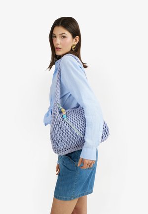 Donna con camicetta azzurro chiaro e gonna di jeans porta una grande borsa a tracolla lavanda intrecciata con perline decorative.