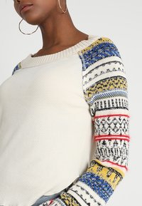 Kermakudisui, jossa on kuvioidut hihat sinisen, keltaisen ja punaisen sävyissä sekä erilaisia geometrisia muotoja. Vartalon tekstuuria ja ribbineuletuin.