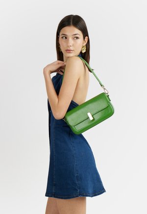 Donna con vestito di jeans blu senza maniche che tiene una borsa a tracolla in pelle verde con fibbia dorata, indossa grandi orecchini dorati, guarda oltre la spalla.