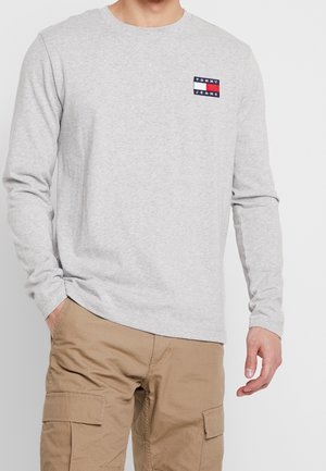 Homme portant une chemise Tommy Jeans à manches longues gris clair et un pantalon cargo kaki, debout avec une main dans la poche sur un fond uni.
