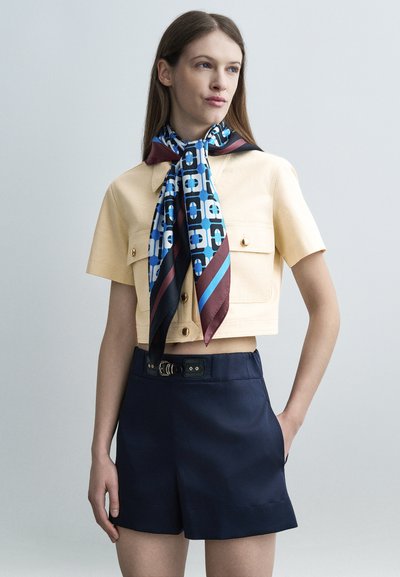 Femme portant une chemise courte beige avec des boutons dorés, un short marine ceinturé et un foulard bleu à motifs noué autour du cou, debout devant un fond gris.