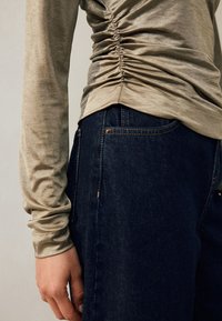 Garām piedurknēm topiņš taupe krāsā ar rievotu detaļu jostasvietā, kombinēts ar tumši ziliem džinsiem ar redzamām šuvēm un kabatu.