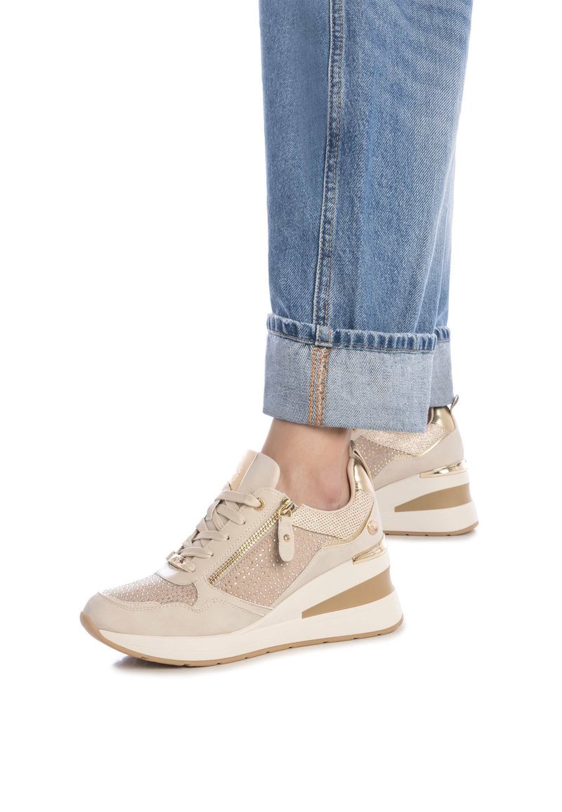 Zapatillas deportivas beige con malla brillante y materiales de ante, que presentan acentos dorados y un diseño de tacón cuña. Tela texturizada y cierre con cremallera.