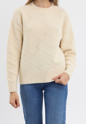 Femme portant un pull en tricot couleur crème avec un motif côtelé diagonal et un jean bleu en denim, debout devant un fond uni.