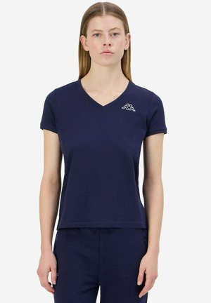 Donna con maglietta a V blu navy con un piccolo logo bianco sul petto a sinistra, abbinata a pantaloni blu navy coordinati.