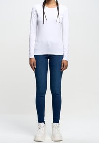 Camisa blanca de manga larga con cuello redondo, combinada con jeans ajustados azul y zapatillas blancas gruesas. Diseño sencillo sin patrones visibles.