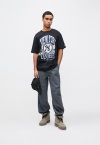 Camisola preta com gráfico "New York Yankees", calças cinzentas largas e botas castanhas. A segurar um boné às riscas preto.