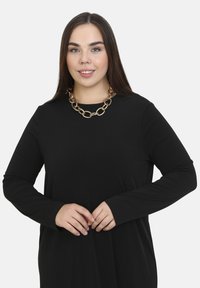Schwarzes langärmliges Kleid mit einer lockeren Passform, das eine runde Ausschnittform und eine große goldene Kette als Akzent bietet.