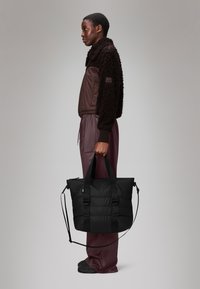 Saco tote acolchoado preto com duas alças grossas e uma alça destacável, posicionado ao lado de uma pessoa vestindo um casaco felpudo castanho e calças combinando.