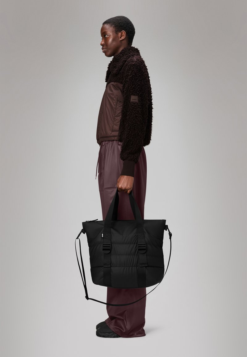 Saco tote acolchoado preto com duas alças grossas e uma alça destacável, posicionado ao lado de uma pessoa vestindo um casaco felpudo castanho e calças combinando.