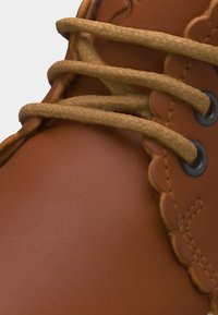 Κοντινό πλάνο καφέ δερμάτινου μποτού με μπεζ στρογγυλά κορδόνια περασμένα από μαύρα μεταλλικά οφθαλμίσκια και δερμάτινη ραφή με κυματιστό σχέδιο.