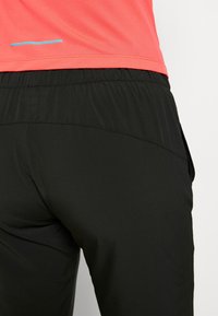 Pantalones deportivos negros con una textura suave, con una cintura elástica y bolsillos laterales. Se ve una camiseta de malla rosa por encima de la cintura.