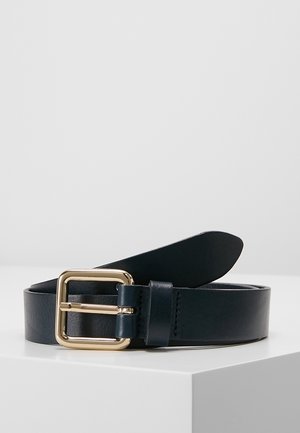 Ceinture en cuir noir avec une boucle carrée dorée enroulée sur une surface blanche sur un fond gris clair uni.