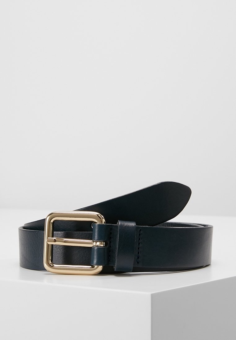 Ceinture en cuir noir avec une boucle carrée dorée enroulée sur une surface blanche sur un fond gris clair uni.