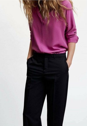 Kobieta ubrana w bluzkę z długim rękawem w kolorze magenta, włożoną do czarnych spodni z wysokim stanem, z rękami w kieszeniach, na jednolitym jasnym tle.