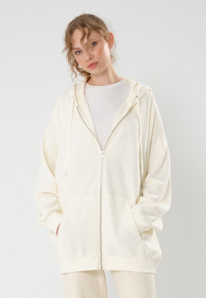 Haut à zipper crème, comprenant une capuche, de grandes poches avant, des poignets côtelés et une coupe décontractée, fabriqué en tissu doux et texturé.