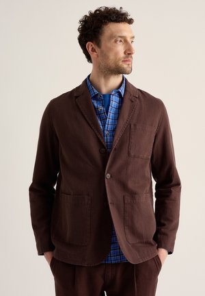 Mann mit lockigem Haar, der einen braunen Blazer über einem blau karierten Hemd trägt, schaut seitlich mit den Händen in den Taschen vor einfachem Hintergrund.