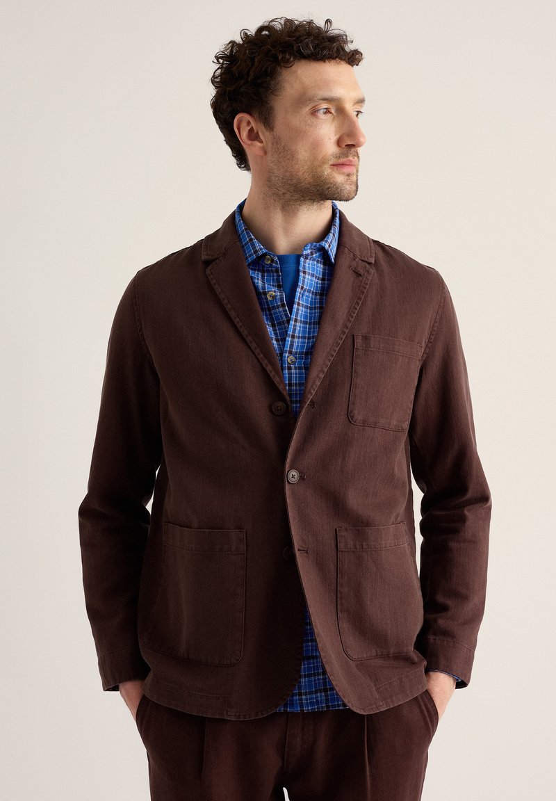 Homme aux cheveux bouclés portant un blazer marron par-dessus une chemise à carreaux bleue, regardant sur le côté, les mains dans les poches, sur un fond uni.