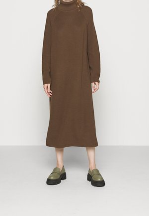 Personne portant une longue robe marron en maille à col roulé et des mocassins épais vert olive, debout sur un sol gris uni et un mur clair.