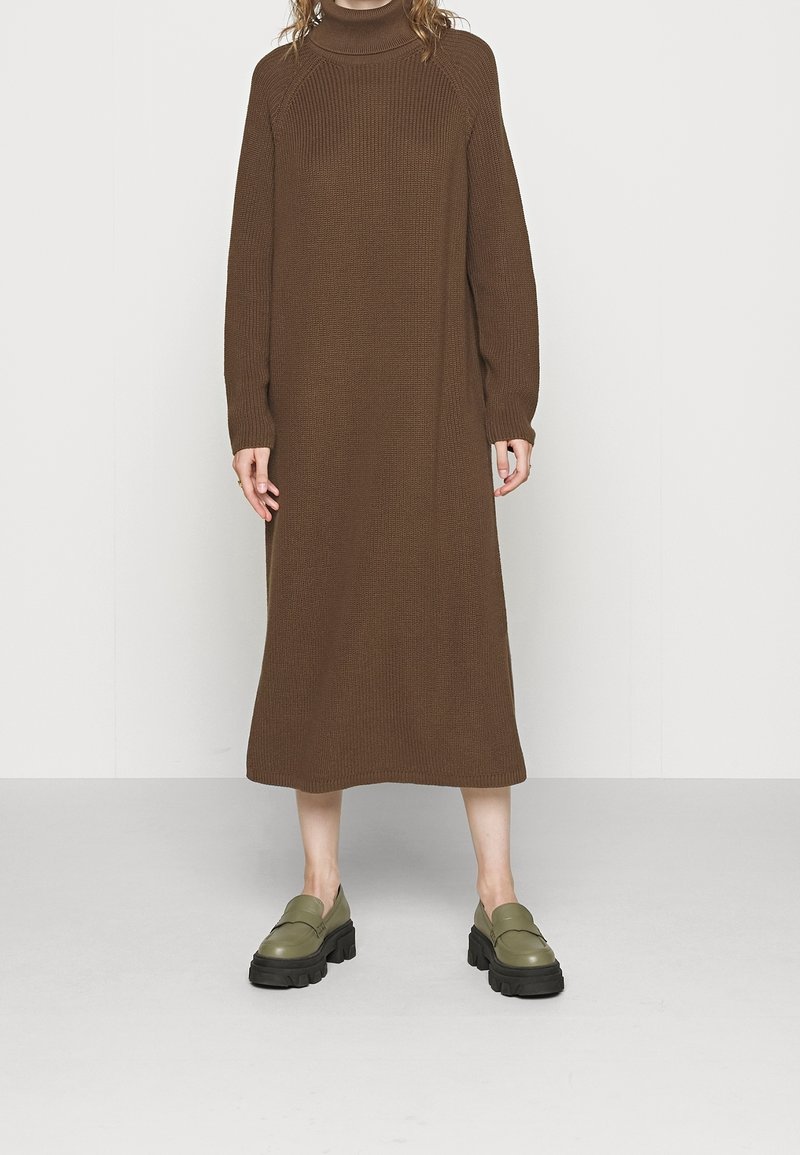 Personne portant une longue robe marron en maille à col roulé et des mocassins épais vert olive, debout sur un sol gris uni et un mur clair.