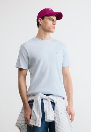 Mladi moški, ki nosi svetlo modro kratko srajco, vijolično kapo, kavbojke in ima okoli pasu zavezano belo srajco z lisami, gleda v desno.