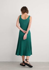 Sleeveless green ribbed dress with a flared pleated skirt. Back view reveals a scoop neckline and crossed arms. Worn with black sandals.