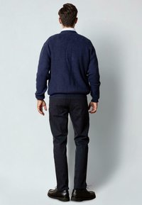 Man staat met zijn gezicht naar een lichtgrijze muur, draagt een marineblauwe trui, zwarte broek, zwarte schoenen en een wit overhemd met kraag.