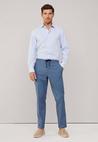 Camicia a quadri azzurro chiaro con bottoni, abbinata a pantaloni scuri con cordino e scarpe beige senza lacci, su uno sfondo neutro.