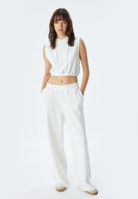 Biały crop top z kapturem i elastycznym dołem w zestawie z luźnymi, białymi spodniami dresowymi. Miękka tkanina i widoczne kieszenie z przodu. W komplecie wygodne obuwie.
