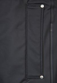Veste en tissu noir au design épuré avec une poche plate et un bouton-pression en argent. Texture lisse avec un minimum de détails de couture.