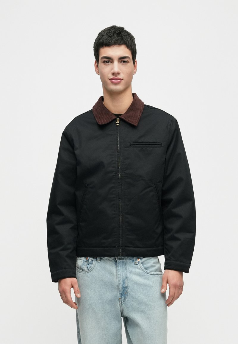 Jaunas vyras dėvi juodą užtrauktuką su rudu koloriumi ir šviesiai mėlynus džinsus, stovintis prieš paprastą baltą foną.