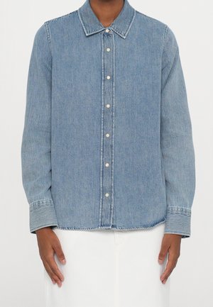 Osoba ubrana w jasnoniebieską, dżinsową koszulę z długim rękawem i białe spodnie, stojąca na tle jednolitej ściany.