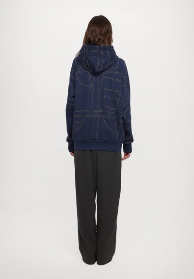 Navy hooded sweatshirt with contrasting beige stitching, featuring a geometric pocket design. Paired with loose-fitting gray sweatpants.