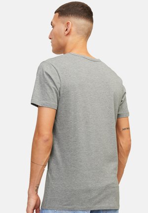 Uomo con capelli corti che indossa una maglietta grigia a maniche corte semplice, visto di spalle su sfondo bianco.