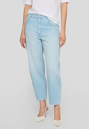Frau trägt hellblaue High-Waist-Jeans, weißes kurzärmeliges Oberteil und weiße Slingback-Pumps mit spitzer Spitze, steht vor einem schlichten Hintergrund.