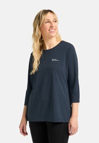 Blonde Frau, die ein navy-blaues Langarmshirt von Jack Wolfskin und schwarze Hosen trägt, leicht nach rechts schaut und lächelt.