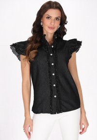 Camicia in denim nero con maniche corte a volant, bottone frontale e dettagli sfrangiati lungo il colletto e la patta dei bottoni. Indossata con pantaloni bianchi.