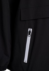 Schwarzer Jackenstoff mit strukturiertem Finish, ausgestattet mit einer weißen Reißverschlusstasche mit schwarzem Ziehband. Enthält eine Klapptasche über dem Reißverschluss.