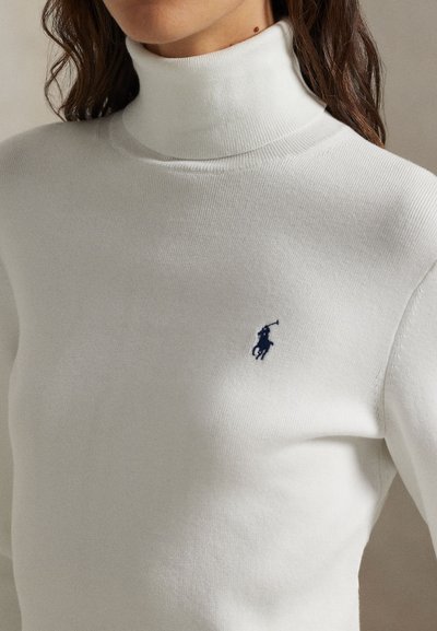 Mujer con un suéter cuello alto blanco ajustado con un pequeño logo de jugador de polo azul marino en el pecho.