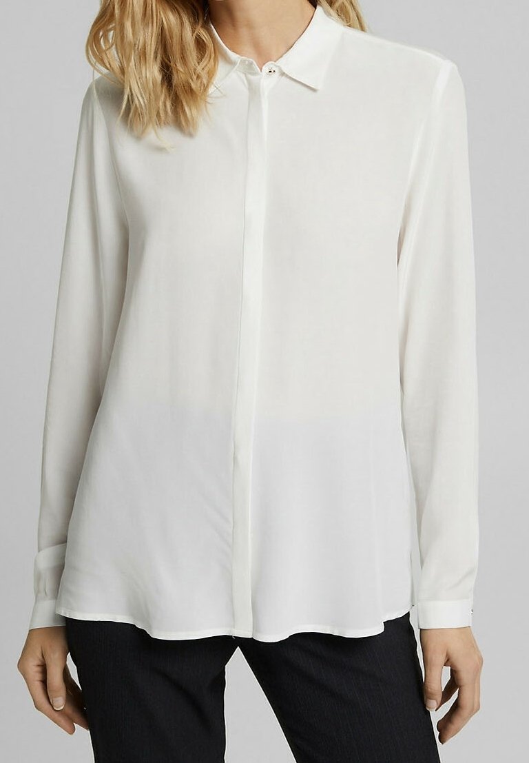 Femme portant un chemisier blanc à manches longues avec col et boutons, associé à un pantalon noir sur fond gris.