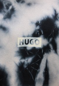Μαύρο και άσπρο πλεκτό ύφασμα με σχέδιο tie-dye, που διαθέτει ένα ορθογώνιο κρεμ ετικέτα με τη λέξη «HUGO» σε έντονα μαύρα γράμματα.