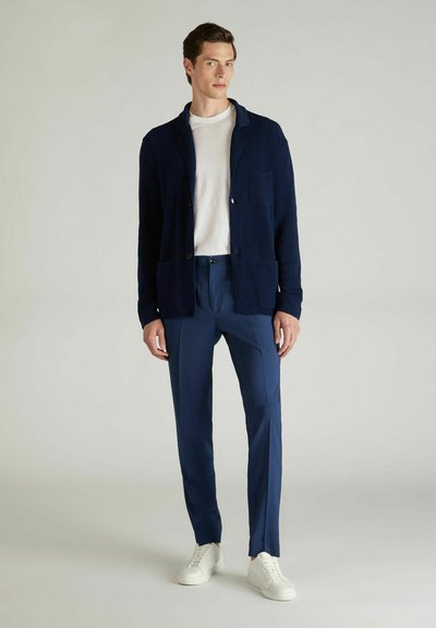 Navy gestrickte Strickjacke mit zwei Fronttaschen, getragen über einem weißen T-Shirt und blauen Hosen, kombiniert mit weißen Sneakers. Einfaches, modernes Design.