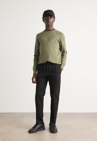 Maglione verde oliva con una piccola toppa, pantaloni neri con cordini e sneakers nere, completati da un cappellino nero. Texture liscia in tutto.