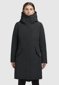 Femme portant un long manteau d'hiver noir avec un col haut et un devant boutonné, se tenant devant un fond gris clair uniforme.