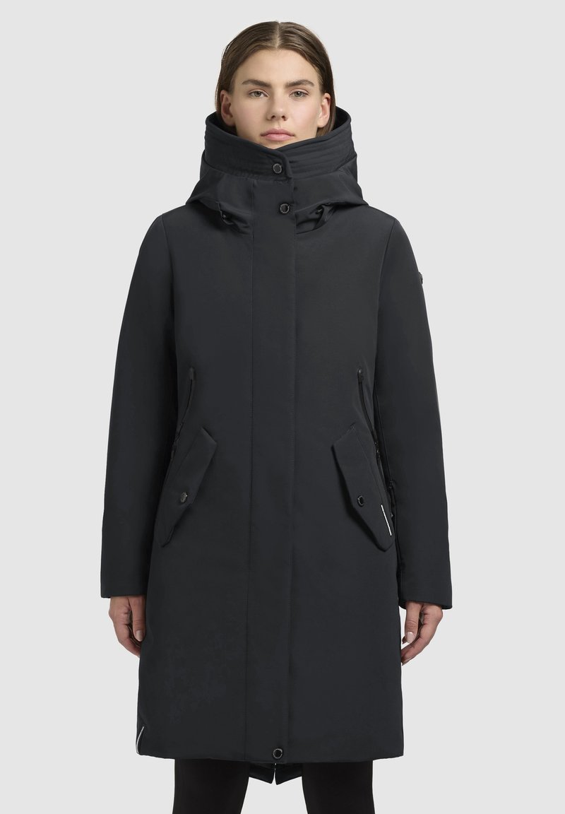 Femme portant un long manteau d'hiver noir avec un col haut et un devant boutonné, se tenant devant un fond gris clair uniforme.