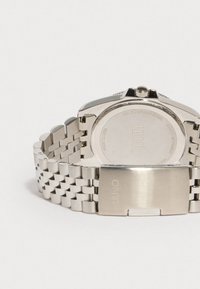 Ruostumattomasta teräksestä valmistettu kello, jossa on metallinen ranneke, pyöreä kuori ja kiinteä takapinta. Kaiverrettu brändilogo ja mallin tiedot näkyvät.