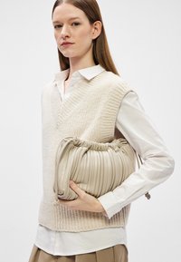 Bolsa de mão de couro plissada em bege com fecho de cordão, segurada debaixo do braço, combinada com um colete de malha texturizado em creme e uma camisa branca.