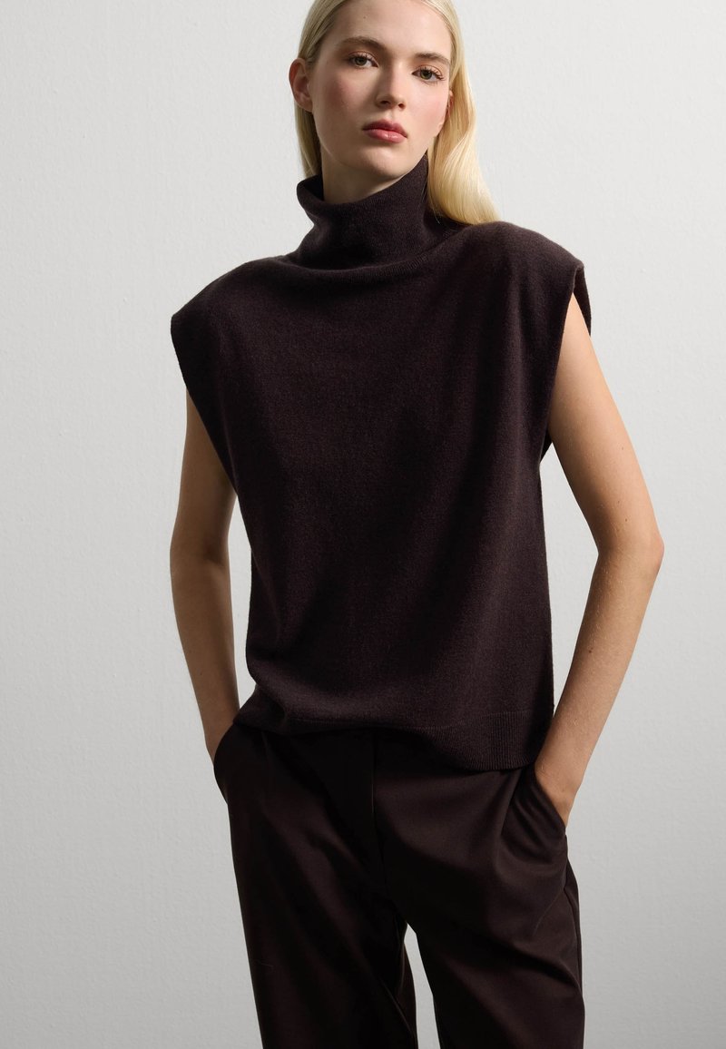 Donkerbruine, mouwloze top met hoge hals van zachte stof; gecombineerd met donkere broek. Honden textuur, minimalistisch ontwerp, casual silhouet.