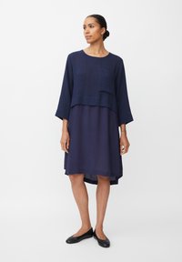 Frau steht in einem dunkelblauen, geschichteten Kleid mit lockerem Oberteil und schwarzen Ballerinas vor einem schlichten weißen Hintergrund.