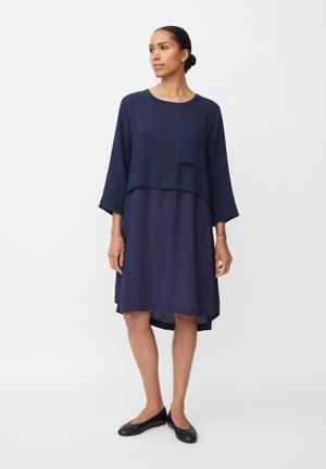Mujer de pie vistiendo un vestido azul oscuro en capas con una parte superior holgada y zapatos planos negros, frente a un fondo blanco liso.
